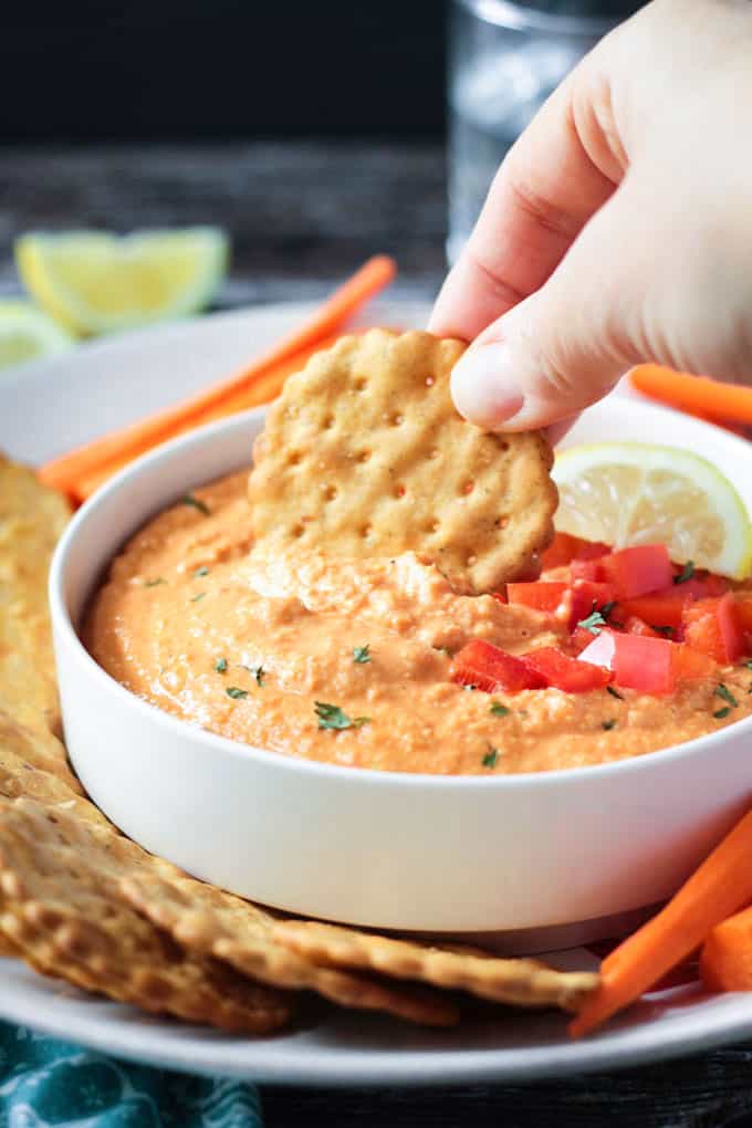 Round cracker being dipped into a bowl of roasted red pepper hummus.