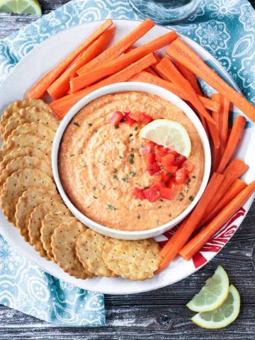 Roasted red pepper hummus in a white bowl on a white plate. Crackers and carrots surround the bowl on the plate.