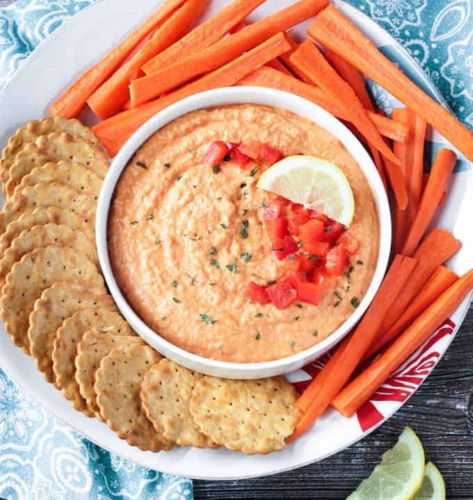 Roasted red pepper hummus in a white bowl on a white plate. Crackers and carrots surround the bowl on the plate.