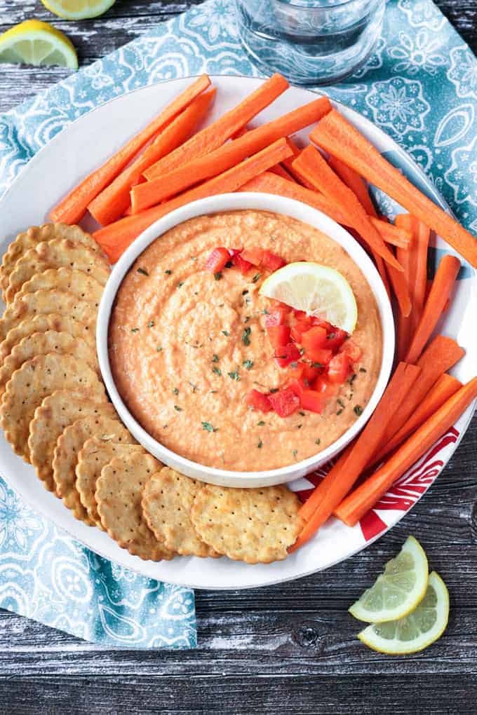 Roasted red pepper hummus in a white bowl on a white plate. Crackers and carrots surround the bowl on the plate. 