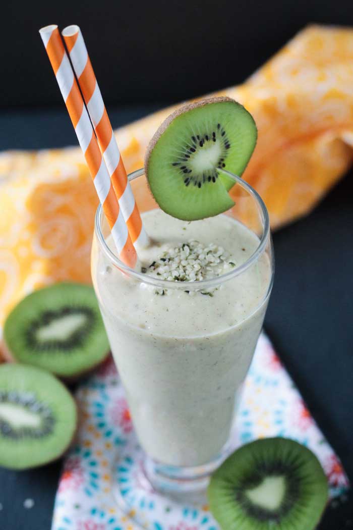 A vibrant green kiwi slice garnishes a tall glass of kiwi mango hemp smoothie with 2 orange and white paper straws.