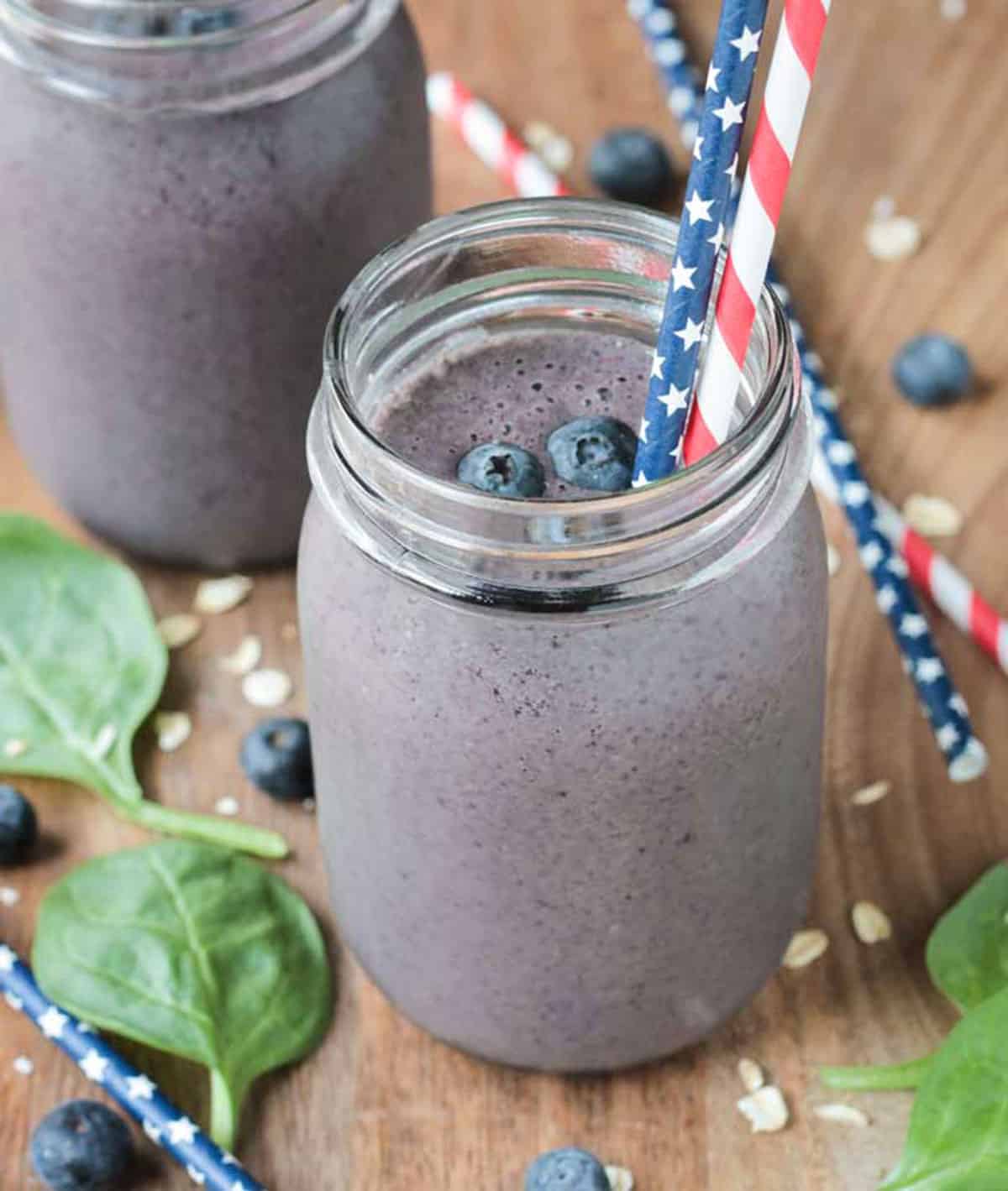 two red, white, and blue straws in a blueberry smoothie surrounded by fresh spinach leaves and fresh blueberries