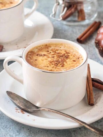 Cup of pumpkin spice coffee on a saucer with a spoon and two cinnamon sticks.