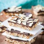 Stack of three Triple Almond Oat Bars separated by squares of parchment paper. More oat bars in the background.