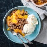 Vegan peach crisp with two scoops of vanilla ice cream on a blue plate.