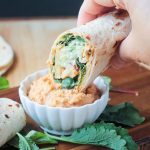 Tortilla veggie wrap dipping into a small white bowl of spicy hummus.