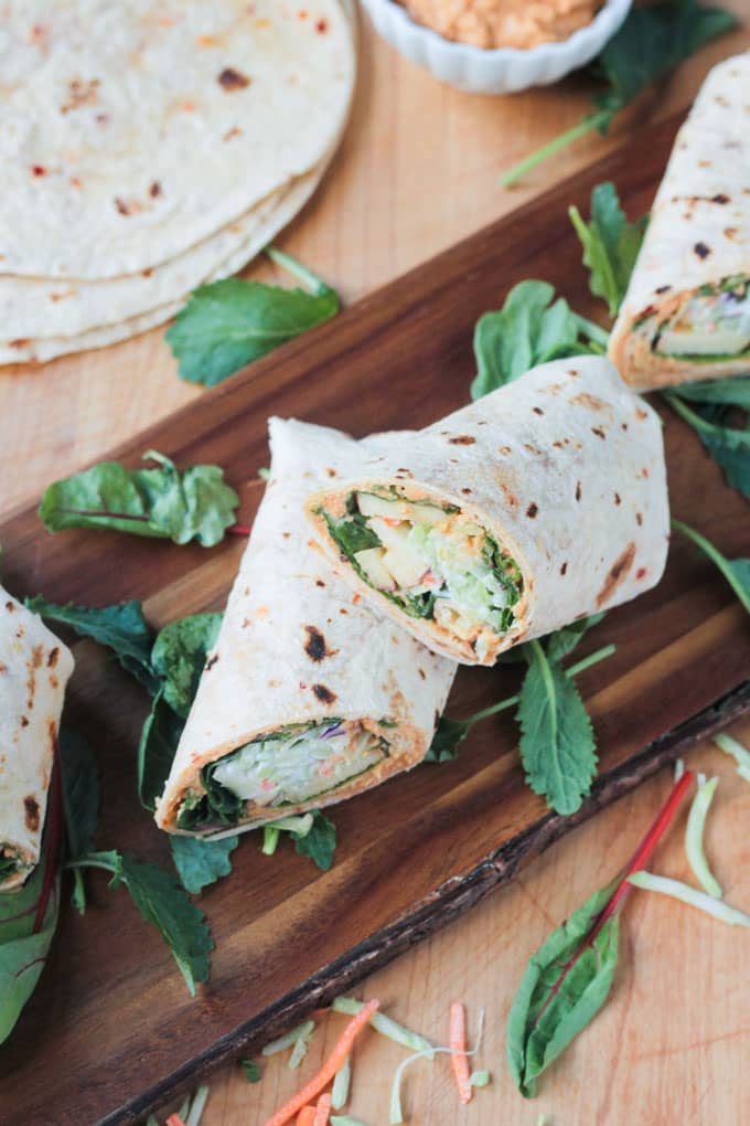 Overhead shot of veggie tortilla wraps on a wood serving board with leafy greens scattered nearby.