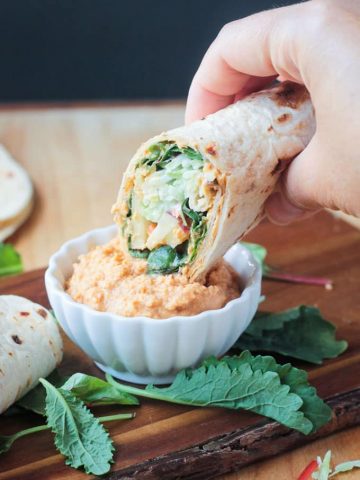 Tortilla veggie wrap dipping into a small white bowl of spicy hummus.