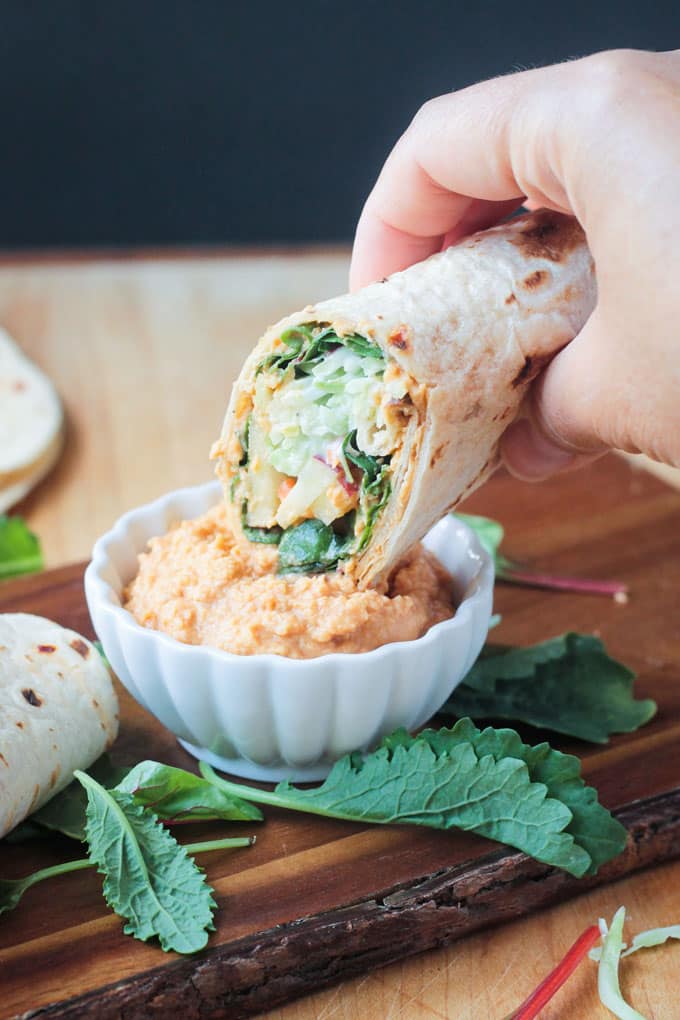 Tortilla veggie wrap dipping into a small white bowl of spicy hummus.