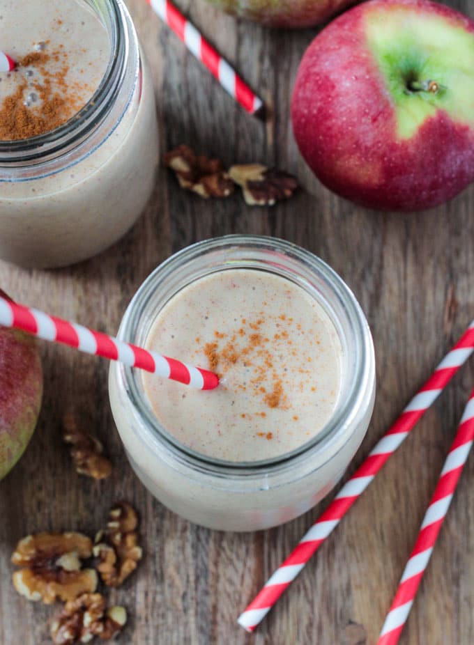 Glass of Apple Pie Smoothie sprinkled with cinnamon with a red and white striped straw in it. Fresh apples and walnuts lying on the table around the glass.