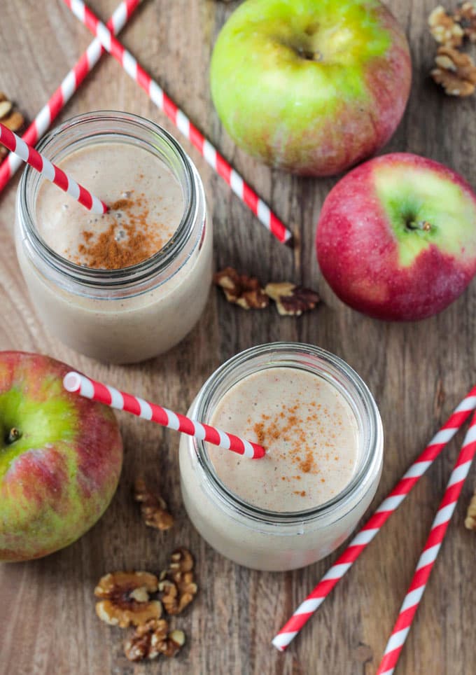 2 glasses of Apple Pie Smoothie with red and white straws. Fresh apples, walnuts, and more straws lie nearby.