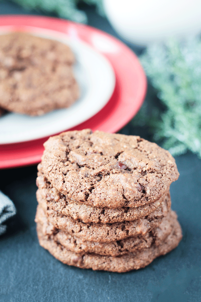 Stack of four Chocolate Almond Cookies in front of a red plate with more cookies.