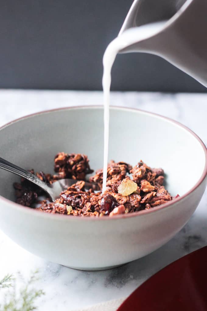 Milk being poured into a rustic stoneware bowl full of granola. A metal spoon rests on the side.
