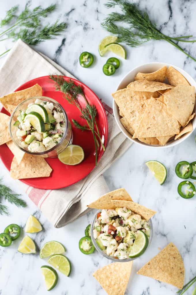 Glass bowl of ceviche on a red plate, another glass bowl of ceviche, and a bowl of tortilla chips all on a marble slab. Jalapeño slices, lime wedges, and fresh dill lie all around.