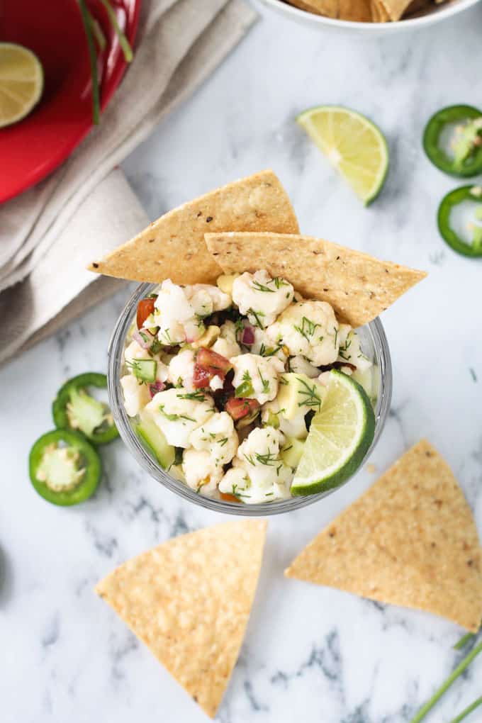 Cauliflower vegan ceviche in a bowl on a marble slab surrounded by tortilla chips and jalapeño slices.