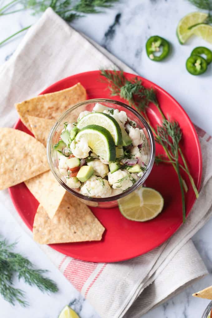 Glass bowl of ceviche, tortilla chips, lime wedge, and fresh dill, all on a red plate.