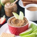 Two apple slices in a bowl of cookie butter next to a cup of coffee and stack of toast.