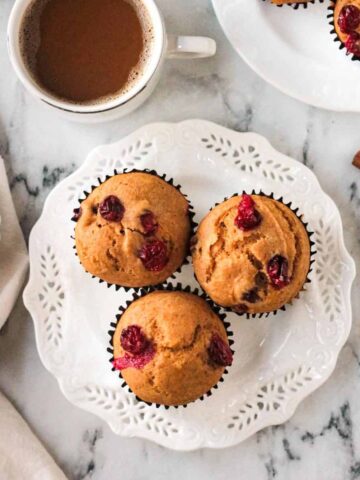 Three pumpkin cranberry muffins on a white plate in front of a cup of coffee.