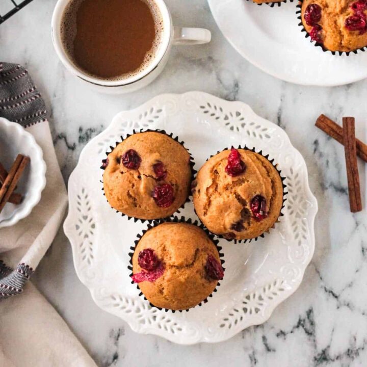 Three pumpkin cranberry muffins on a white plate in front of a cup of coffee.