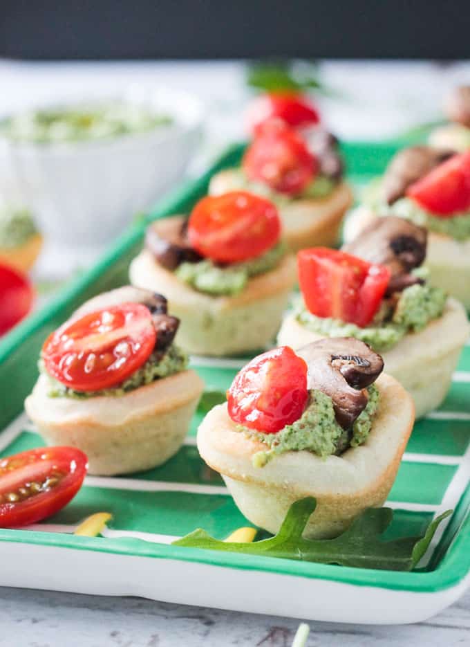 Vegan Pizza Bites - easy pizza dough topped with dairy free pesto, mushrooms and cherry tomatoes. Big bold flavors in a little bite - perfect for your next party or get together! Especially great for game day! Close up of vegan pizza bites on a green tray. Small bowl of pesto in the background.