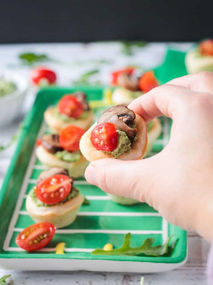 Hand holding one vegan pizza bite. Tray full of more pizza bites in the background.