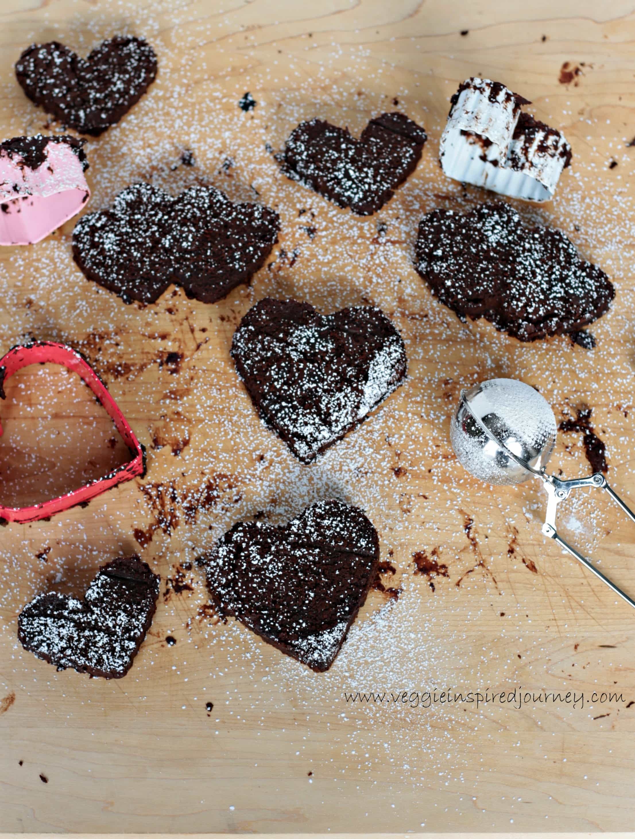Overhead view of a batch of Fudgy Mocha Black Bean Brownie Hearts next to heart shaped cookie cutters and a small mesh tea ball infuser used to sprinkle powdered sugar.