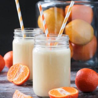 Two dairy free creamy clementine smoothies in front of a large glass container of oranges.