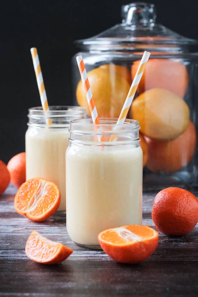 Two dairy free creamy orange banana smoothies in front of a large glass container of oranges.