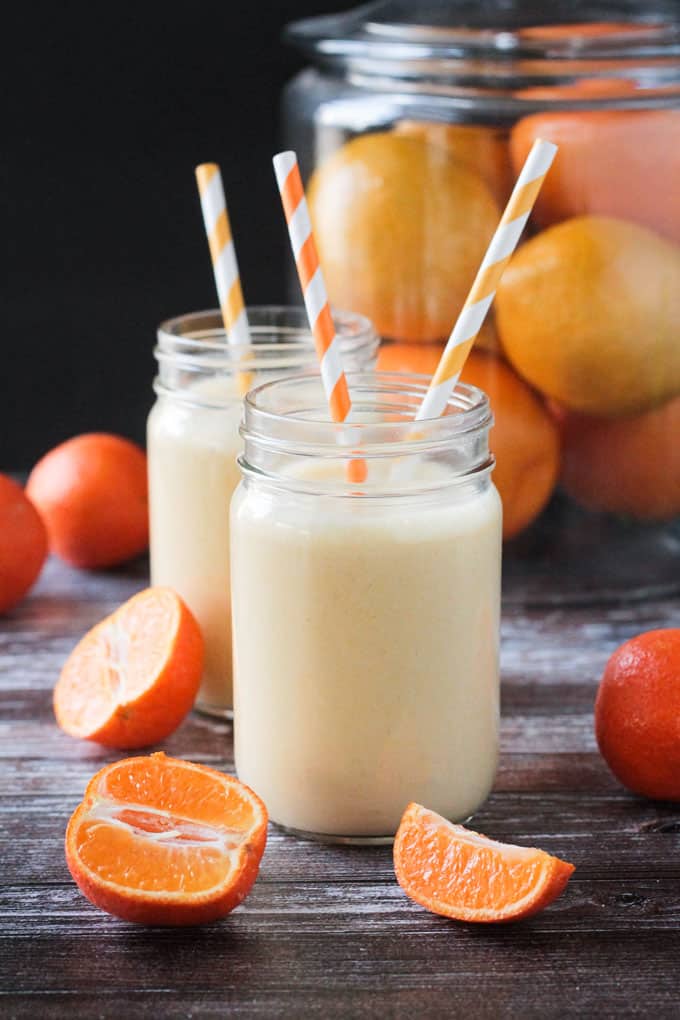 Two glass of orange smoothie wit orange/white straws. Halved clementines rest nearby.