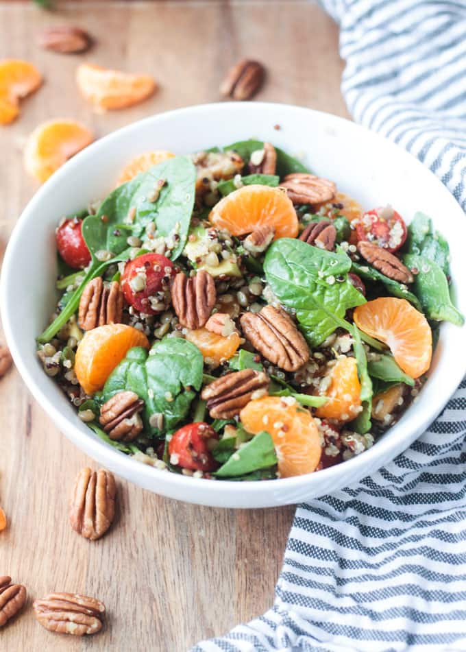 Salad of spinach, orange slices, tomatoes, quinoa, lentils, & pecans in a white bowl next to a gray and white striped dish towel. 