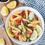 Overhead shot of a bowl of penne pasta w/ vegetables & garnished with lemon slices. A gray & white striped dish towel lies next to the bowl on the right. A fork, lemon wedges, and a small bowl of olives is on the left side of the bowl.