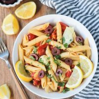 Overhead shot of a bowl of penne pasta w/ vegetables & garnished with lemon slices. A gray & white striped dish towel lies next to the bowl on the right. A fork, lemon wedges, and a small bowl of olives is on the left side of the bowl.