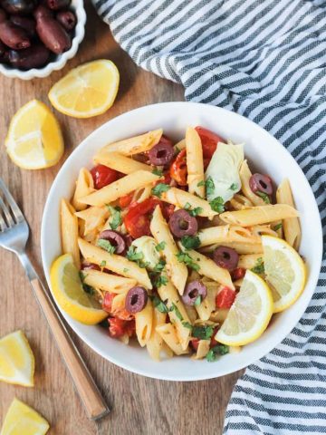 Overhead shot of a bowl of penne pasta w/ vegetables & garnished with lemon slices. A gray & white striped dish towel lies next to the bowl on the right. A fork, lemon wedges, and a small bowl of olives is on the left side of the bowl.