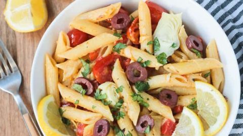 Overhead shot of a bowl of penne pasta w/ vegetables & garnished with lemon slices. A gray & white striped dish towel lies next to the bowl on the right. A fork, lemon wedges, and a small bowl of olives is on the left side of the bowl.