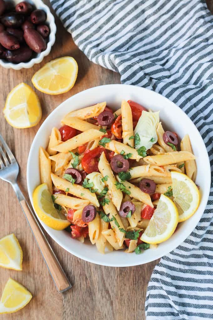 Overhead shot of a bowl of penne pasta w/ vegetables & garnished with lemon slices. A gray & white striped dish towel lies next to the bowl on the right. A fork, lemon wedges, and a small bowl of olives is on the left side of the bowl.