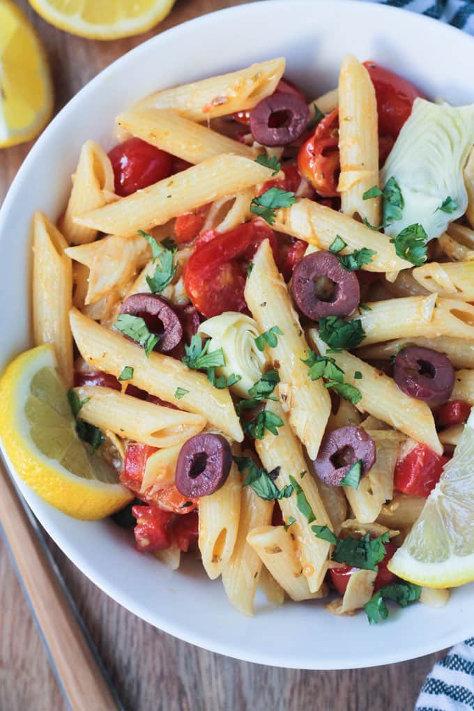 Close up overhead shot of a bowl of penne pasta w/ roasted red peppers, cherry tomatoes, artichoke hearts, and olives. Garnished with chopped parsley and lemon slices.