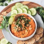Roasted carrot hummus in a bowl with crackers and cucumbers on the side.