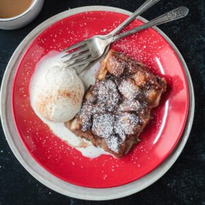 Slice of vegan bread pudding on a red plate with a scoop of vanilla ice cream.