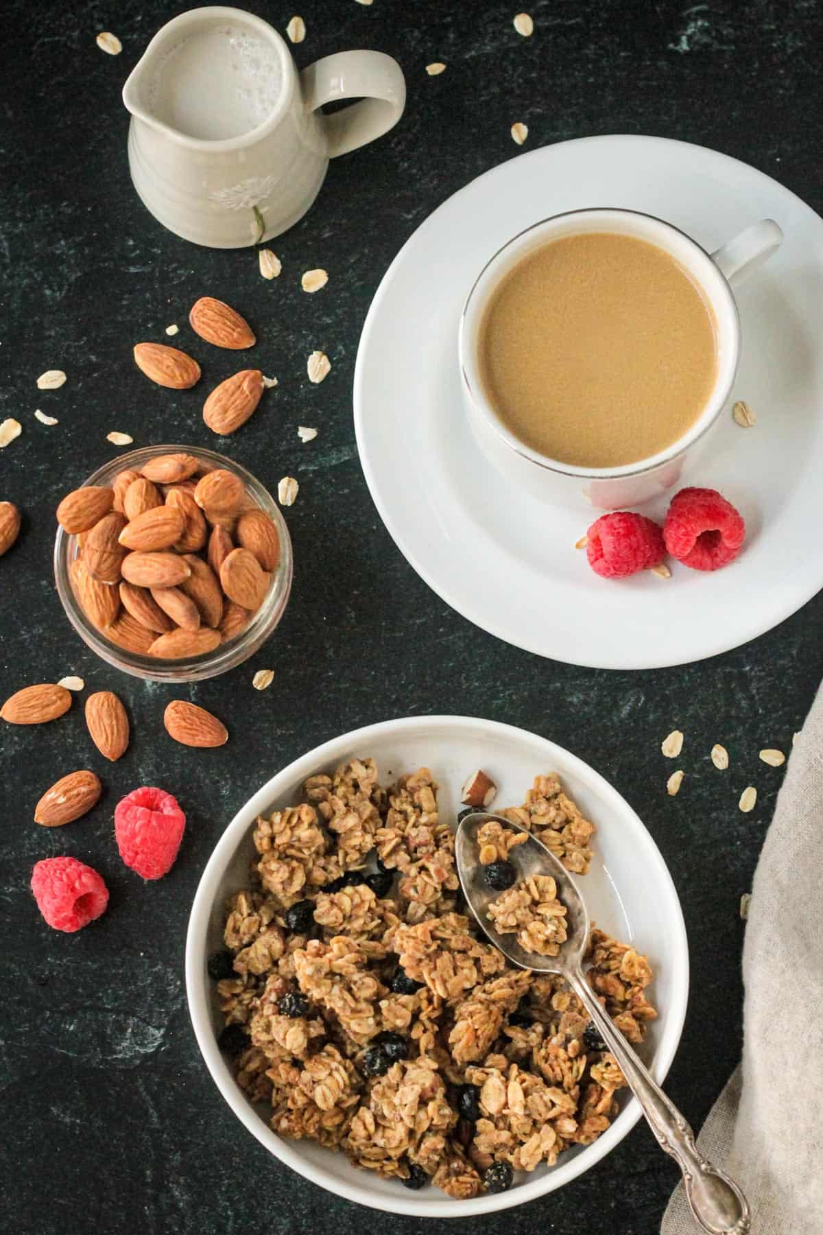 Bowl of almond granola next to a cup of coffee and a small jar of almonds.