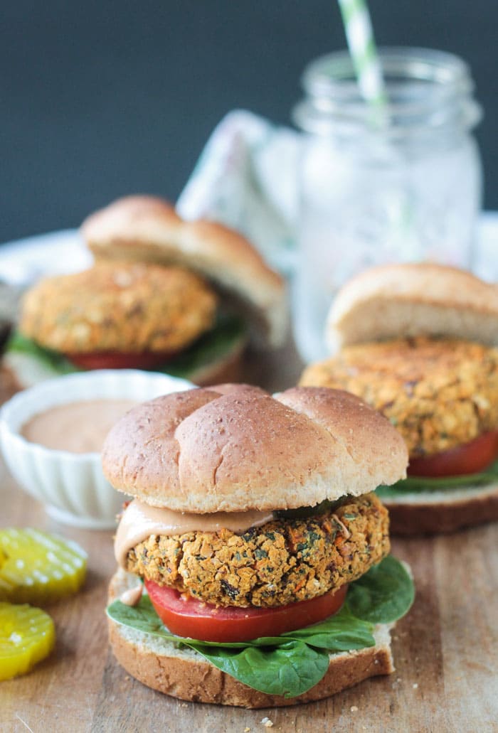 Veggie burger on a bun with spinach leaves, a tomato slice, and bbq sauce. Pickle slices and a small bowl of extra bbq sauce on the side. Two more burgers on buns in the background.