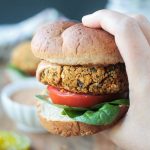 Close up shot of a hand holding a veggie burger on a wheat bun with spinach leaves, a tomato slice, and bbq sauce.