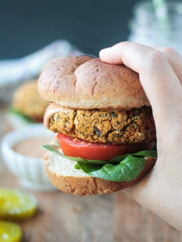 Close up shot of a hand holding a veggie burger on a wheat bun with spinach leaves, a tomato slice, and bbq sauce.