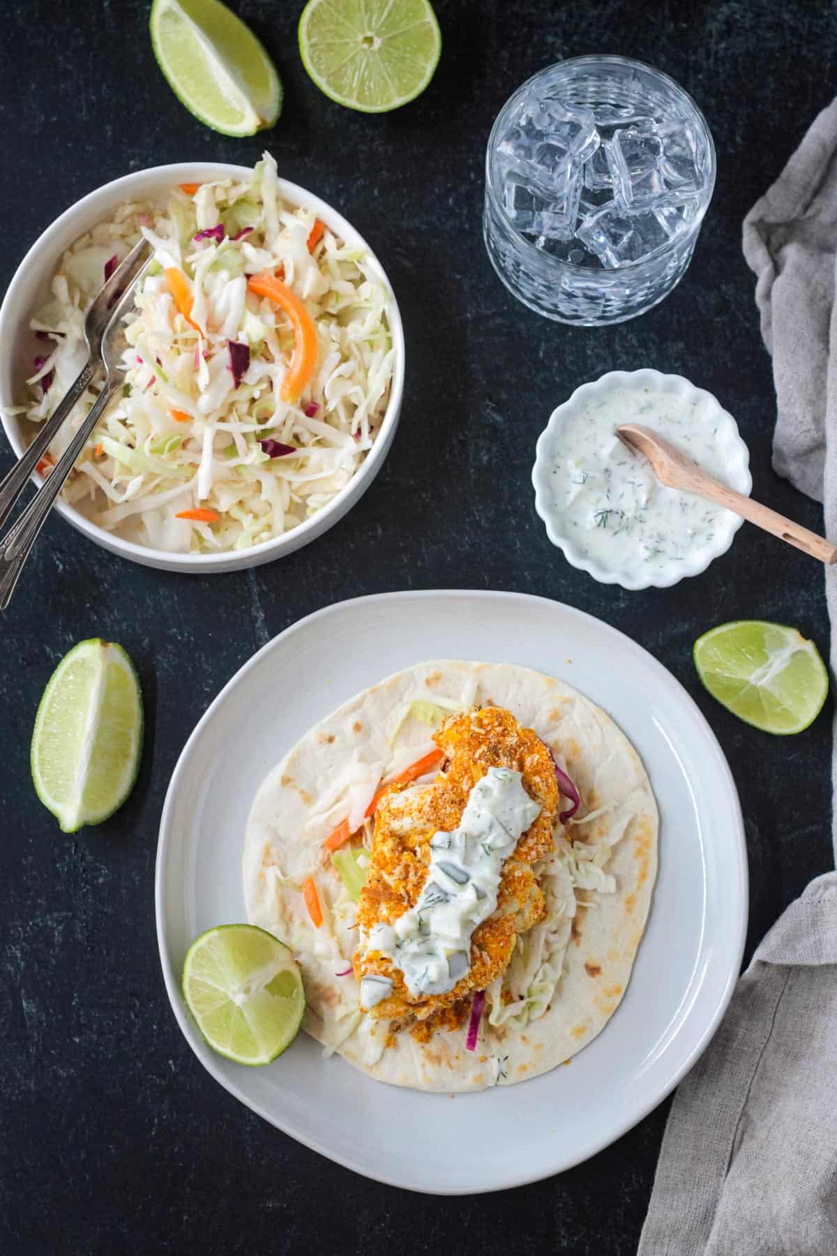 Tablescape with one cauliflower taco on a plate, bowl of coleslaw, bowl of tartar sauce, glass of water, and fresh lime wedges scattered about.