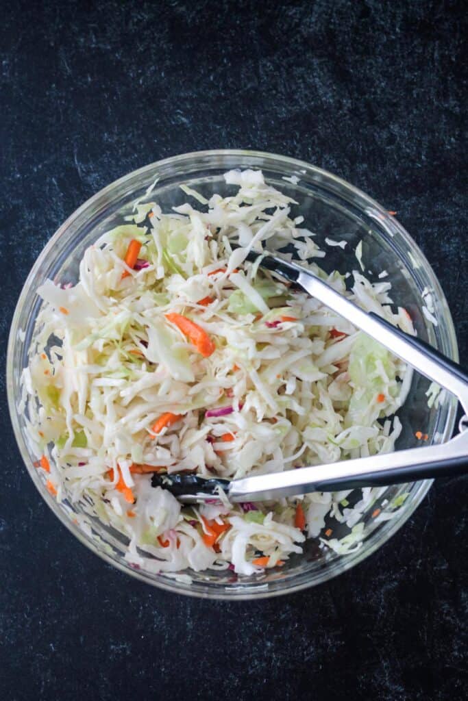 Coleslaw in a glass bowl with serving tongs.