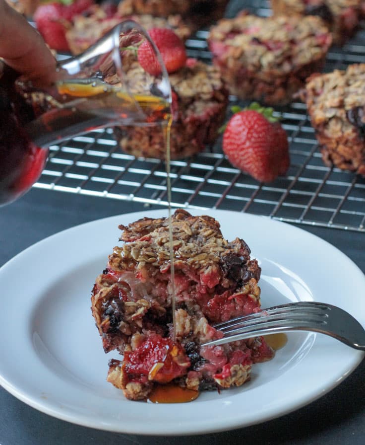 A silver fork cutting open one Strawberry Banana Baked Oatmeal muffin as pure maple syrup is drizzled from a small glass jar above.