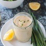 Vegan ranch dressing in a glass jar on a white plate next to chives and a lemon wedge.