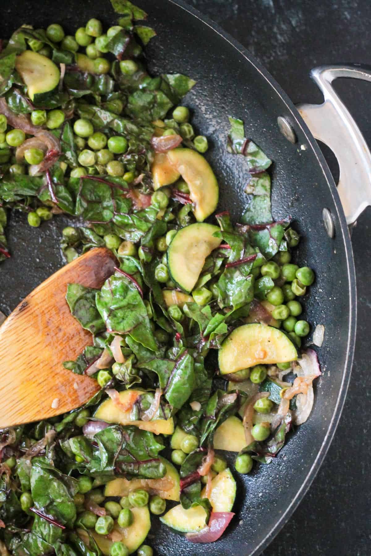 Wooden spoon stirring sautéed vegetables in a skillet.