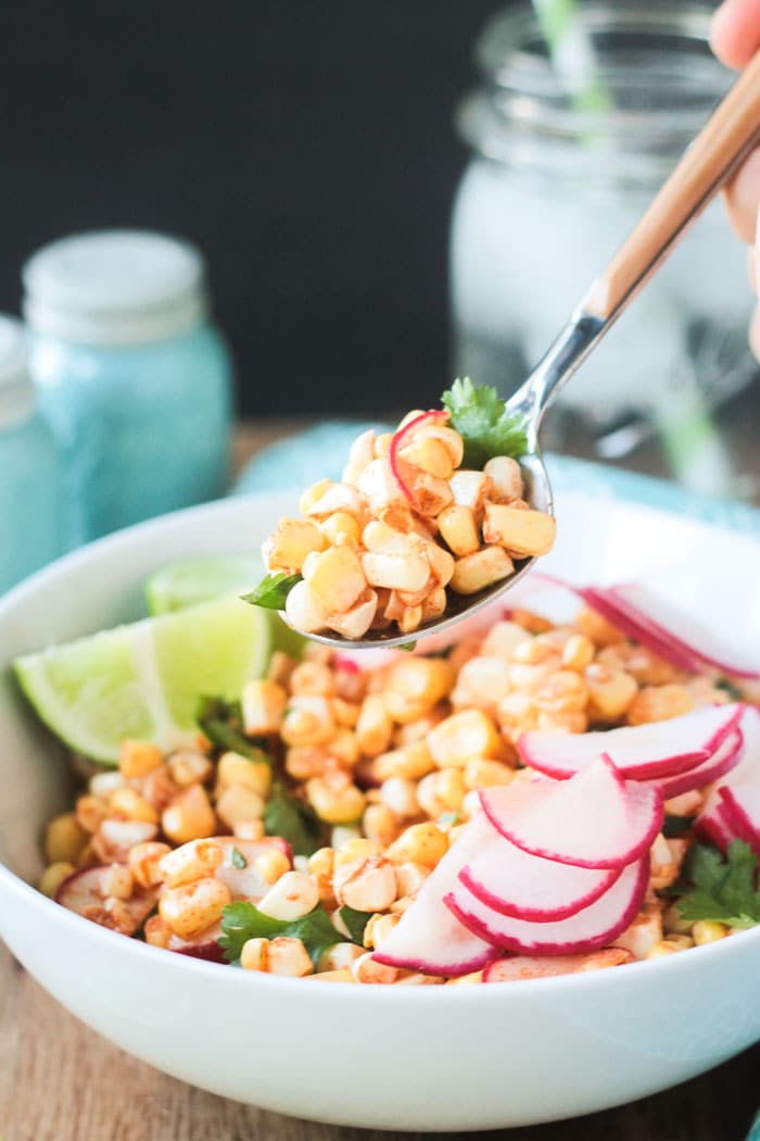 Spoonful of corn and radishes being lifted out of a white bowl containing Raw Corn and Radish Salad.