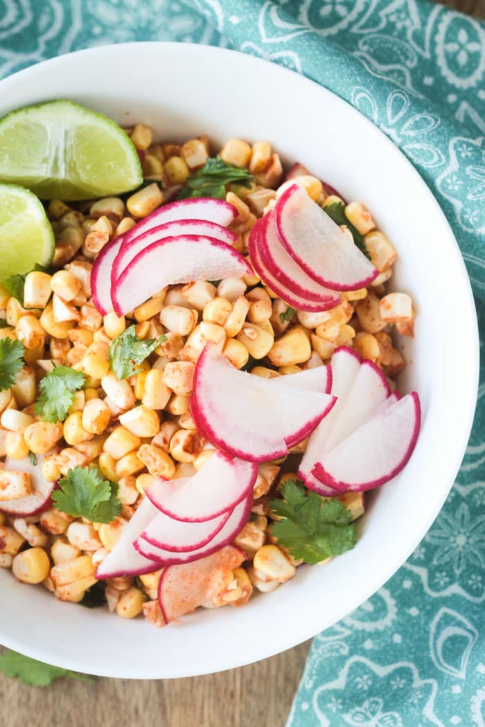 Salad of raw corn and radishes with cilantro and lime wedges in a white bowl.