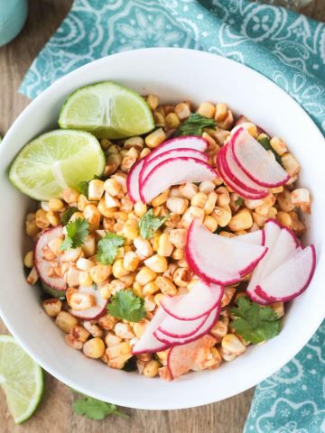 Raw Corn Radish Salad in a white bowl garnished with extra thinly sliced radishes, lime wedges, and cilantro leaves.